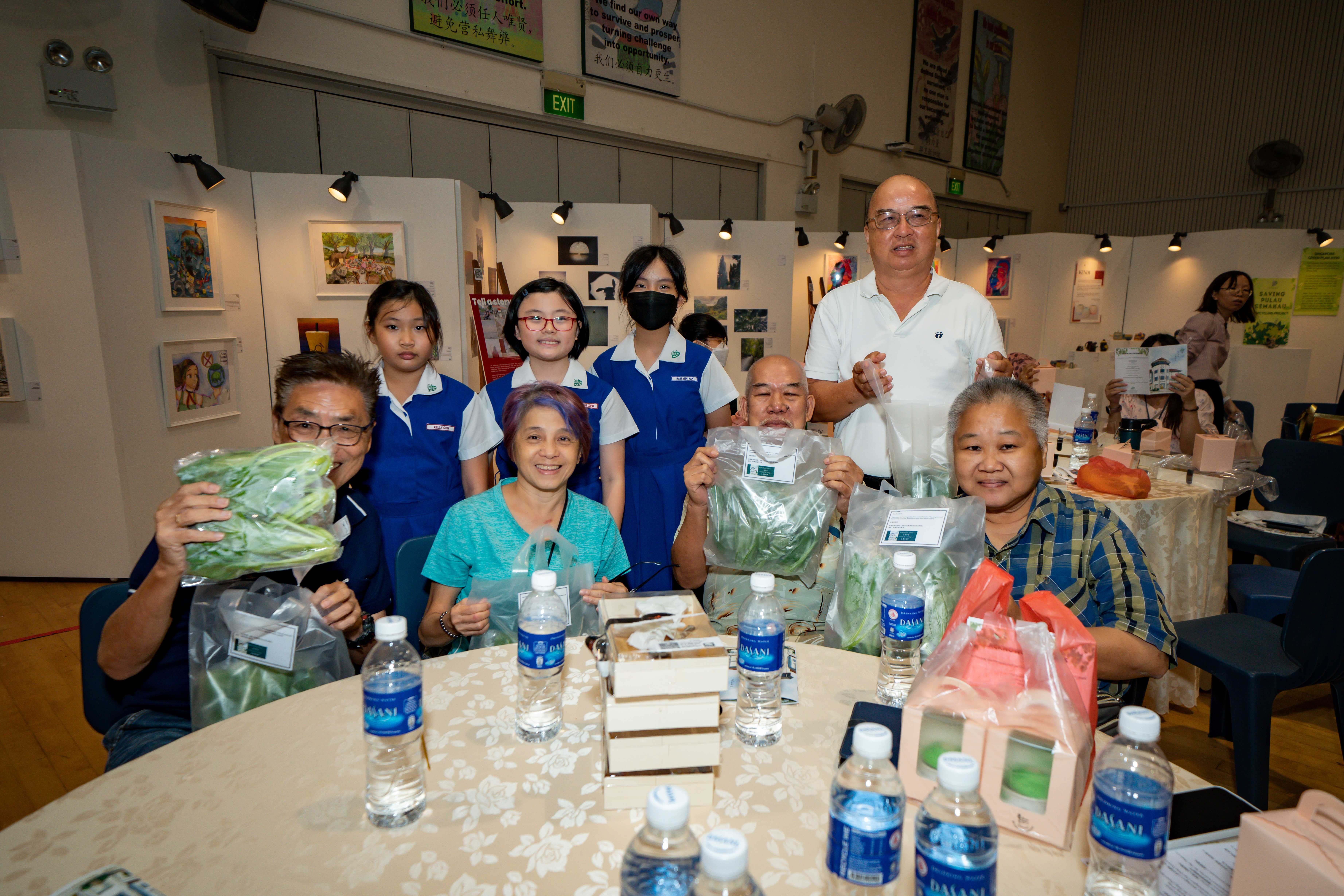 Group of adults and children in blue uniforms holding bags of vegetables at a table with water bottles indoors.